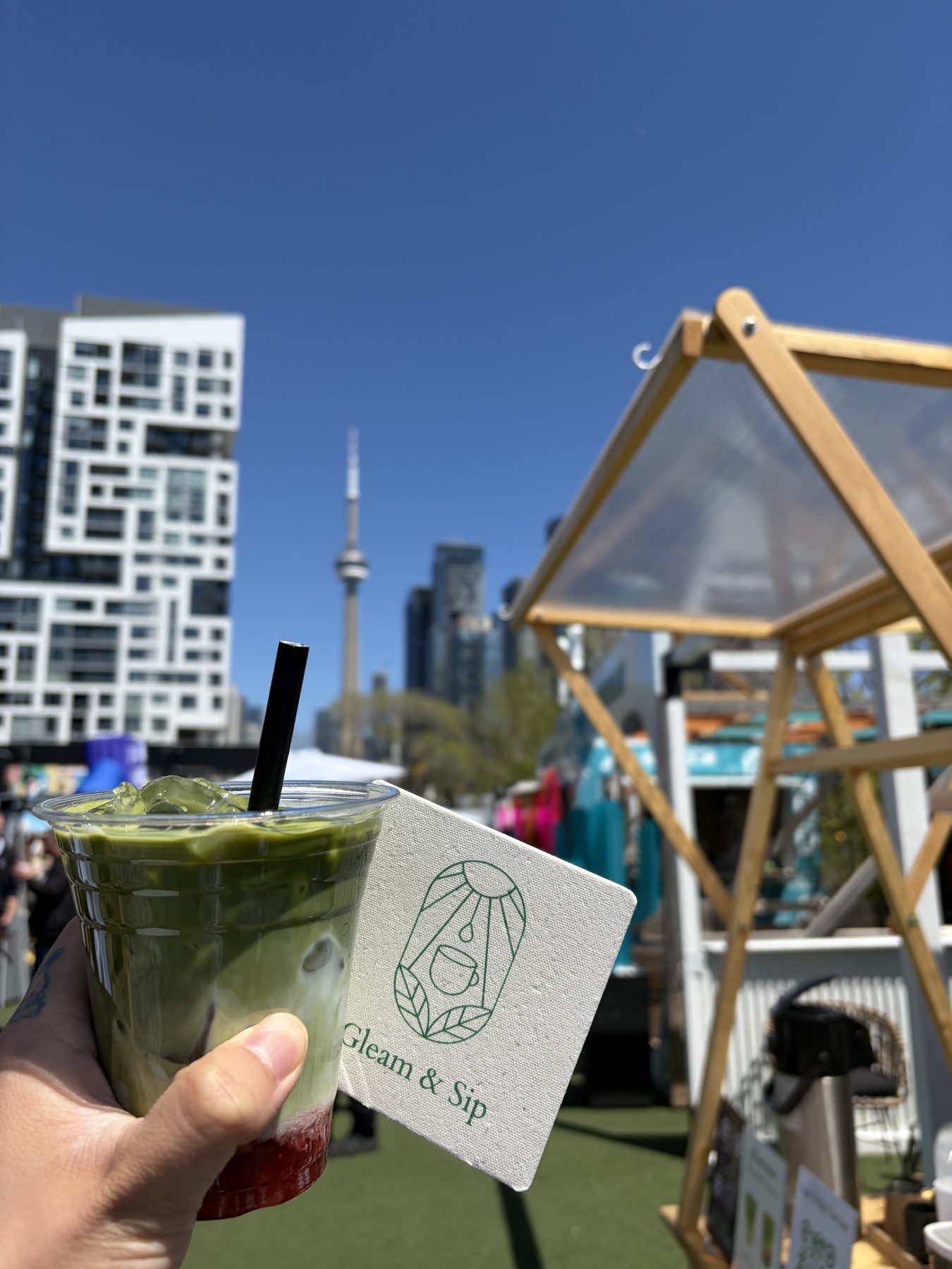 An iced ceremonial matcha with a Gleam & Sip linen coaster held up against the Toronto skyline and CN Tower at Native Touch's AD Digital Day, Stackt Market